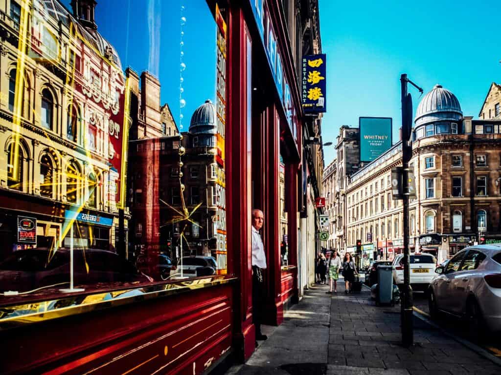 A photo of a British high street. In the shop window is a reflection of other shops on the other side of the road including a Greggs bakery.